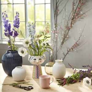 This image contains a variety of vases in different shapes and colors displayed on a wooden table, with colorful flowers arranged in some of them, near a window allowing natural light. A whimsical Wiinblad studio vase with its distinctive smiling face adds a playful touch to the setup.