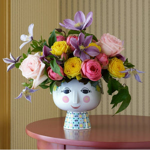 This image features a whimsical vase with a smiling face design, filled with an arrangement of colorful flowers. The flowers include what appears to be roses and clematis, among others, in shades of pink, purple, and yellow. The vase is placed on a pink-toned surface, possibly a table, and is set against a striped beige and brown background, likely a wall. The playful design of the vase, coupled with the vibrant flowers, lends a cheerful and decorative element to the setting.