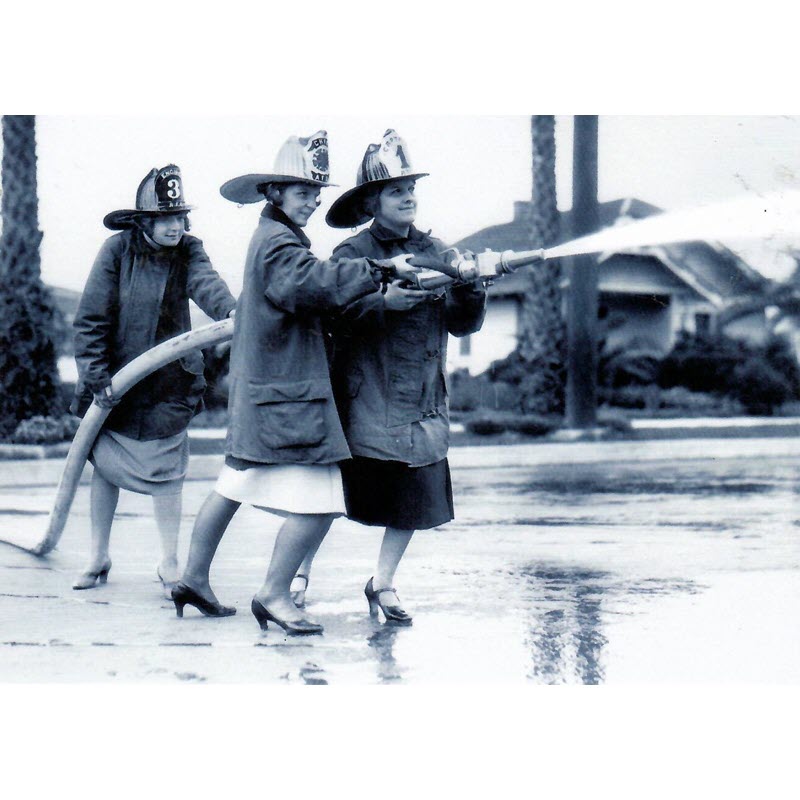 Front of card that has three women in heels with raincoats and firemen's hats holding a fire hose spirting out water.