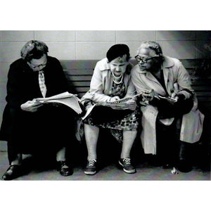 Three older woman sitting on a bench hunched over reading the newspaper. 